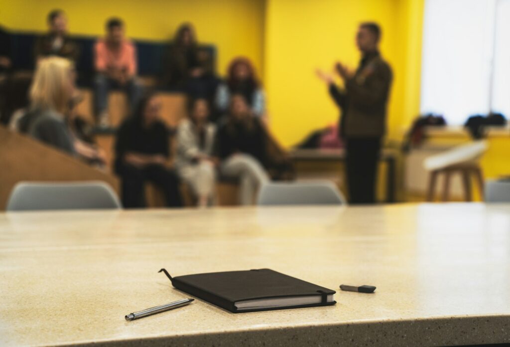 Notebook and pens on a table with a speaker and audience blurred behind, suggesting a training session