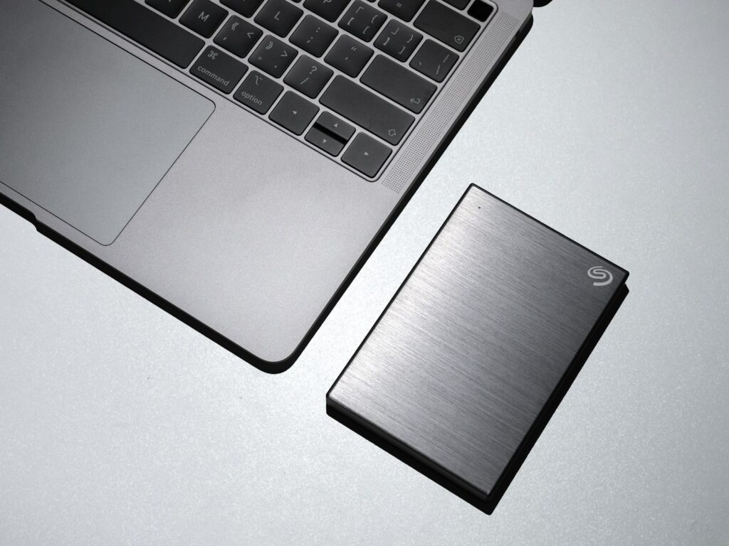 Brushed stainless steel external hard drive beside a MacBook, representing a local backup copy of files